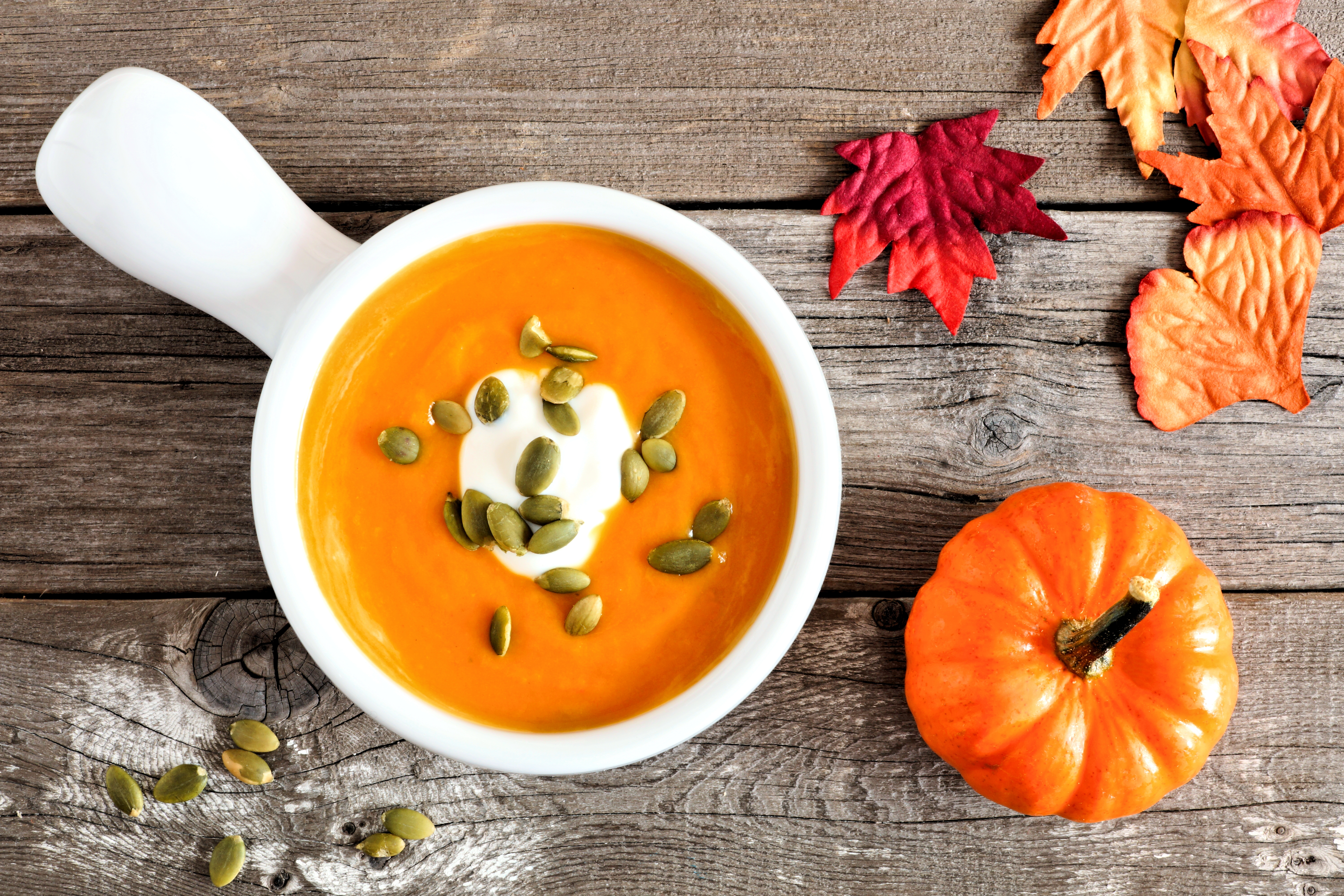 Creamy pumpkin soup topped with pumpkin seeds and cream on rustic wood background with autumn leaves